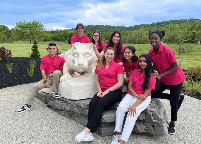 8 Penn State students in pink New Student Orientation shirts are gathered around the Penn State Lion Shrine statue.